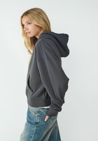 Szara bluza z kapturem w miękkim materiale, z przednią kieszenią i ściagaczami u dołu. Posiada pojemny kaptur oraz luźny krój, zestawiona z niebieskimi dżinsami.