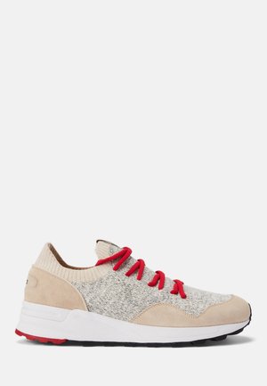 Baskets basses avec tige en maille beige et en suède, lacets rouges, semelle blanche, et semelle extérieure noire et rouge sur fond uni.