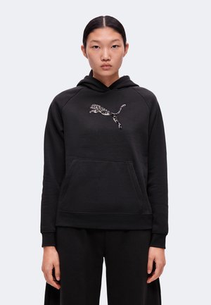 Giovane con capelli neri legati indossa una felpa con cappuccio nera con il logo Puma argentato lucido sul petto, in piedi davanti a uno sfondo semplice.
