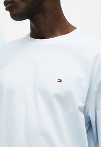 Άνδρας που φορεί ένα απλό λευκό T-shirt με στρογγυλή λαιμόκοψη και ένα μικρό ορθογώνιο κόκκινο, άσπρο και μπλε λογότυπο στο αριστερό στήθος.