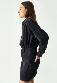 Mekko mustasta denimpistä, jossa on pitkät hihat, korkea kaulus, etutaskut ja vartalonmyötäinen vyötärö. Teksturoitu kangas klassisella muotoilulla.