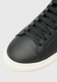 Adidași din piele neagră cu o parte superioară texturată, având șireturi negre și o talpă din cauciuc alb. Vârf rotunjit și detalii minime de cusătură.