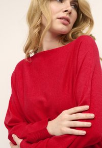 Maglione rosso in lana con scollo a barca e maniche lunghe. Il tessuto ha una consistenza liscia e i polsini presentano dettagli a coste.