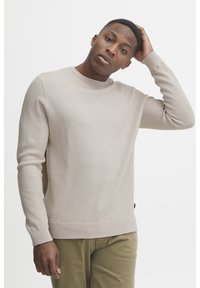 Beige strukturstickad tröja med rundad krage och ribbade ärmmuddar. Bärs över olivgröna byxor med ett subtilt mönster och slät yta.