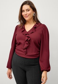 Burgunderfarbene Bluse mit Rüschenausschnitt und langen Puffärmeln, strukturierten Stoff, kombiniert mit schwarzen Hosen, die einen dezenten Glanz haben.