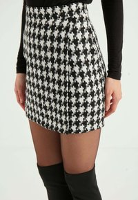 Gonna mini houndstooth bianco e nero realizzata in tessuto texturizzato, con vita aderente e pences laterali. Indossata con stivali neri sopra il ginocchio.