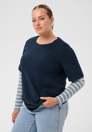 Femme aux cheveux tirés en arrière portant un t-shirt bleu marine par-dessus une chemise à manches longues rayée et un jean bleu clair, posant avec une main dans la poche.