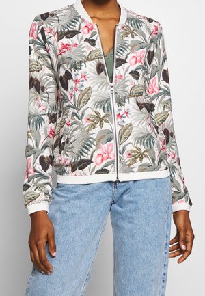 Personne portant une veste blanche zippée avec des motifs floraux et feuillus multicolores, assortie à un jean denim bleu clair.