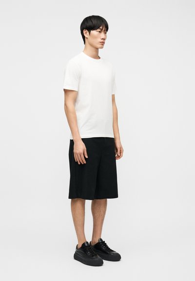 Νέος άνδρας που στέκεται, φορώντας ένα απλό λευκό κοντομάνικο μπλουζάκι, μαύρες βερμούδες μέχρι το γόνατο και μαύρα αθλητικά παπούτσια, με φόντο ένα λευκό τοίχο.