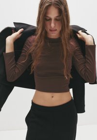 Braunes langärmeliges Crop-Top aus glattem Stoff, kombiniert mit hochtaillierten schwarzen Hosen. Eine schwarze Jacke liegt über einer Schulter.