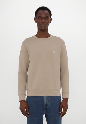 Felpa beige a girocollo con maniche lunghe, polsini e orlo a coste; presenta un piccolo logo sul petto. Abbinata a jeans blu.