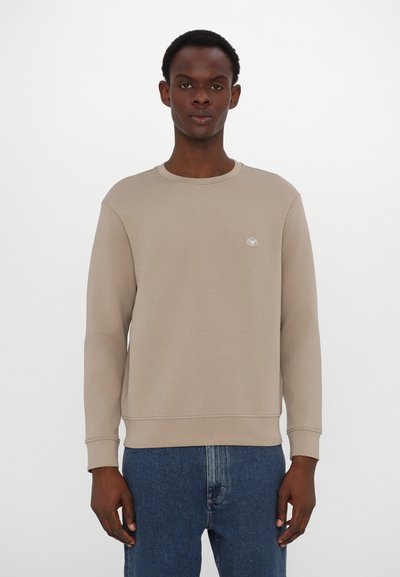 Felpa beige a girocollo con maniche lunghe, polsini e orlo a coste; presenta un piccolo logo sul petto. Abbinata a jeans blu.