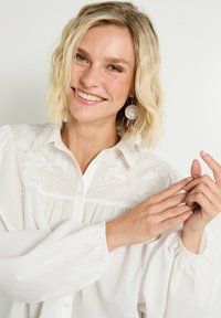 Blusa blanca con cuello y detalles bordados en el canesú, mangas abullonadas y corte holgado. Un par de pendientes circulares añade contraste.