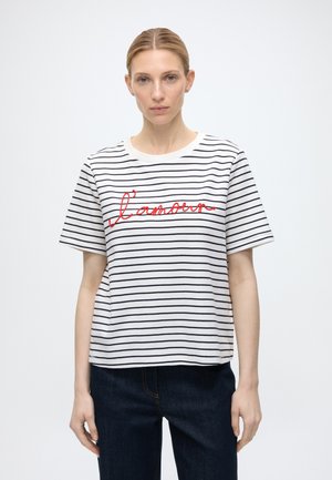 Frau mit zurückgebundenem hellem Haar trägt ein weißes kurzärmeliges Shirt mit schwarzen horizontalen Streifen und rotem Schriftzug "l'amour," kombiniert mit dunkler Hose.