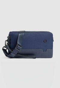 Cartera azul marino con tela texturizada y panel inferior liso, que cuenta con cierre de cremallera y correa ajustable. Detalle de logo destacado en el frente.