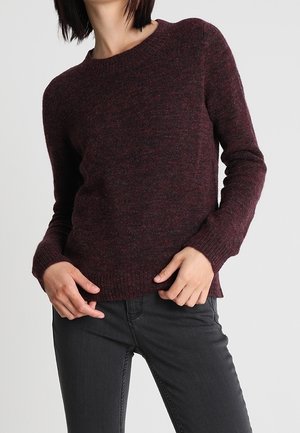 Vrouw draagt een donker bordeauxrode gebreide trui en zwarte broek, met handen ter hoogte van de taille tegen een effen lichte achtergrond.