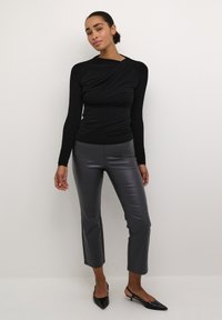 Schwarzes Langarmoberteil mit verdrehtem Vorderdesign, kombiniert mit enganliegenden grauen Kunstlederhosen und spitzen schwarzen Absätzen. Glatte Textur.