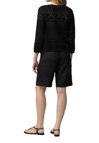 Schwarzes gehäkeltes Oberteil mit Dreiviertelärmeln, kombiniert mit schwarzen Leinen-Shorts. Die Sandalen haben einen einfachen Knöchelriemen und eine Zehenlasche.