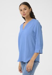 Blouse bleu clair à encolure en V avec des manches trois-quarts, en tissu texturé, détails de fente sur les côtés et coupe légèrement décontractée. Assortie à un jean.