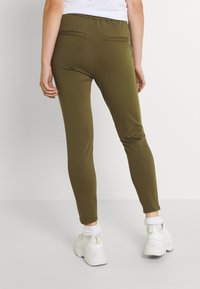 Leggings elásticas verde-oliva com uma textura suave, apresentando uma largura de elástico na cintura e dois bolsos traseiros. Usados com ténis brancos.