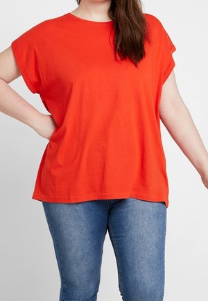 Kvinde iført en løstsiddende, lyse-orange T-shirt med korte ærmer kombineret med tætsiddende blå cowboybukser, poserer foran en ensfarvet baggrund.