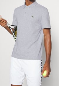 Szara koszulka polo o fakturowanym wykończeniu, z dwoma guzikami przy kołnierzyku i małym zielonym logo. Noszona z białymi szortami; trzyma rakietę i piłkę tenisową.