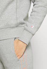 Sudadera de algodón gris con puños acanalados, que presenta un logo en la manga y texto naranja brillante a lo largo del lateral de pantalones de chándal a juego.