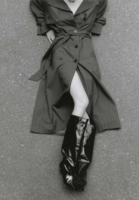 Svartvitt bild som visar en modell i en randig omslagsklänning med en slits och glänsande svarta knähöga stövlar, vilket framhäver släta texturer och djärva kontraster.