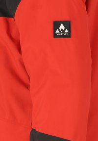 Rode en zwarte jas met een gladde textuur, voorzien van een vierkant logopatch op de bovenarm. Het ontwerp heeft contrasterende kleuren en minimale detailing.