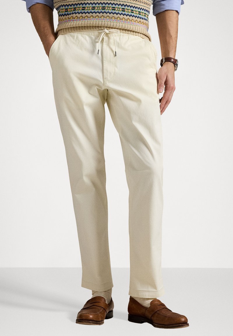 Pantaloni din bumbac de un bej deschis, cu talie elastică, șnur și buzunare laterale, purtați cu pantofi din piele maro închis.