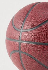 Zbliżenie na fakturowaną czerwoną piłkę do koszykówki, pokazującą czarne szwy i szczegóły powierzchni z ziarnistą fakturą, na jasnym tle.