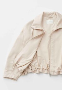 Giacca beige chiaro con zip, collo, cintura elastica e polsino con coulisse regolabile, distesa su una superficie bianca.