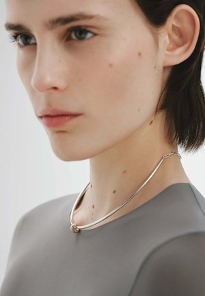 Collar de metal plateado con un acento circular dorado, que presenta un diseño suave y curvado. Lucido sobre una blusa gris, enfatizando el escote.