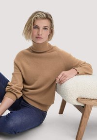 Helles braunes, gestricktes Rollkragensweatshirt mit gerippten Bündchen, cropped Design und weicher Textur. Kombiniert mit dunklen Jeans an einem flauschigen Hocker.