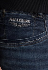 Temno modri jeans iz denima z vezenim logotipom "PME LEGEND" in obliko krila na zadnjem žepu ter kontrastnim rjavim šivom.