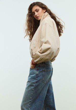 Beige Übergröße-Jacke mit hohem Kragen, hängenden Schultern und großen Taschen. Kombiniert mit lockeren, ausgewaschenen blauen Jeans. Glatte Stoffstruktur.
