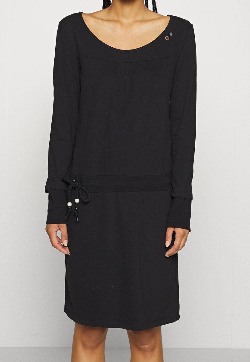 Femme portant une robe noire à manches longues avec un col rond et un détail cordon à la taille gauche.