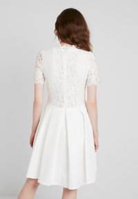 Robe blanche avec un corsage en dentelle, des manches courtes et une jupe plissée, fermeture éclair au dos et texture de tissu lisse.