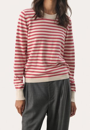 Femme portant un pull à manches longues rayé rouge et blanc avec des poignets côtelés et un pantalon plissé gris, debout devant un fond uni.