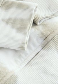 Rippstoff in hellbeige und weiß mit einer strukturierten Oberfläche und unregelmäßigem Verblassungsmuster, ausgestattet mit einem gefalteten Saumdetails.