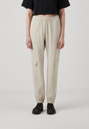 Beige cargo-style pants with an elastic waist and cuffs, featuring two front pockets and a smooth texture. Worn with black footwear.