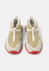 Chaussures slip-on beige et blanches avec semelle en caoutchouc rouge, dotées d'une fermeture éclair et d'un dessus en tissu texturé. Design léger avec un col rembourré.