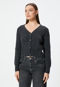 Donkergrijze geribde cardigan met een V-hals en vijf knopen. Heeft lange mouwen en een cropped zoom. Gecombineerd met hoge zwarte jeans.