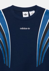 Bluză adidas bleumarin cu dungi curbate albastre, negre și albe pe umeri, având logo-ul Adidas în centrul pieptului.
