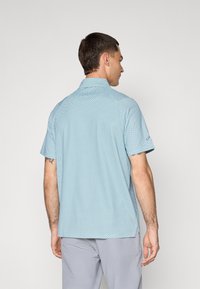 Short-sleeve polo shirt in light blue with a geometric pattern, made of a textured fabric. Features a collar and side vents.