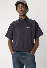 Polo de manga curta, em azul marinho escuro, com colarinho abotoado. Apresenta mangas raglan em contraste e um logo branco no peito.