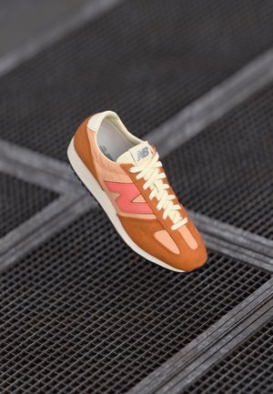 Baskets New Balance orange et rose avec lacets et semelle blancs, posées sur une surface en grille métallique sombre.