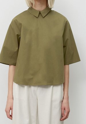 Persoon die een olijfgroen overhemd met korte mouwen en een kraag draagt, gecombineerd met losse witte broek, tegen een effen lichte achtergrond.