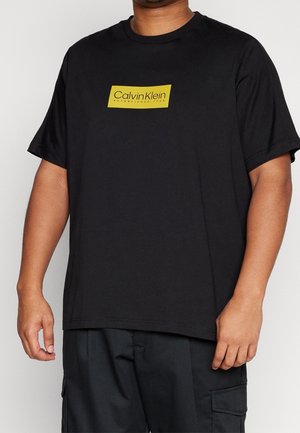 T-shirt nera in cotone con un logo rettangolare giallo che riporta "Calvin Klein Established 1908". Maniche corte e colletto a giro.