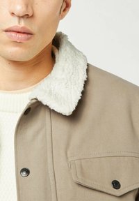 Giacca beige con un colletto morbido bianco, dotata di un design semplice, chiusure a bottoni automatici e una tasca sul petto con un bottone.